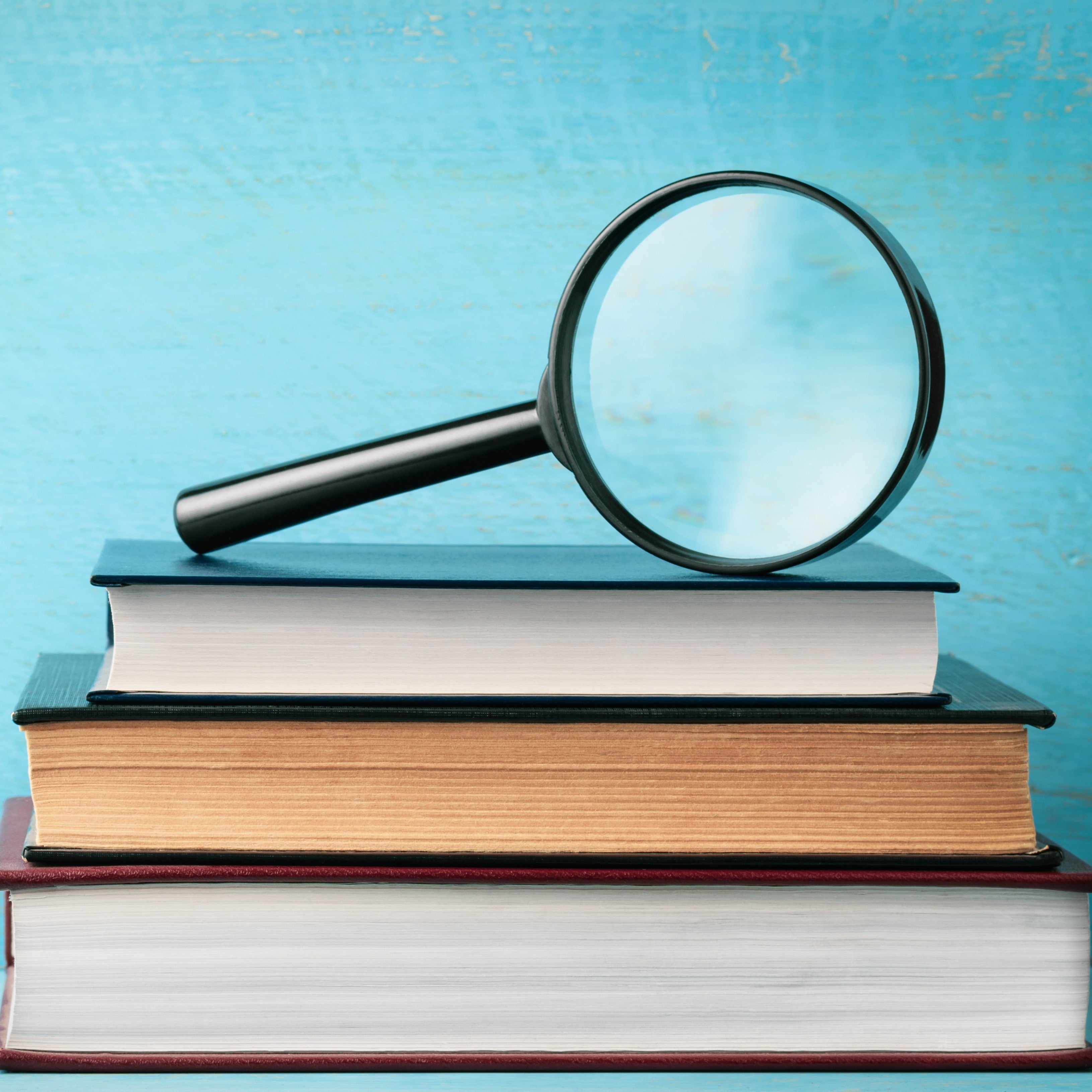 <Black handle magnifying glass sitting on top of three hardback dictionaries with a blue background. Credit> AdobeStock_253293677