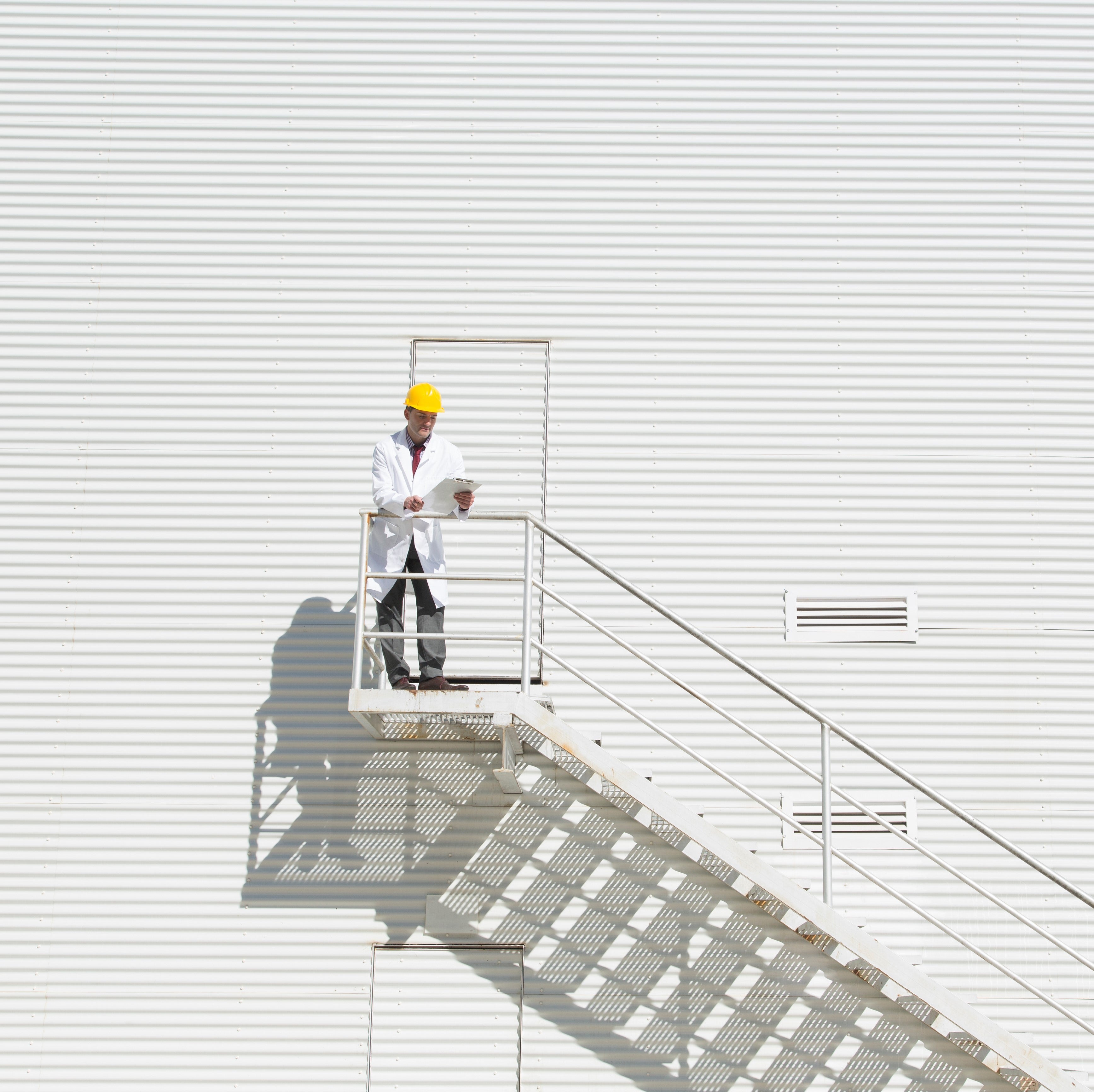 <Employee in white coat and yellow hard hat stood outside a door at height half way up the metal white wall of a building with a stairwell down. Credit> AdobeStock_383183290