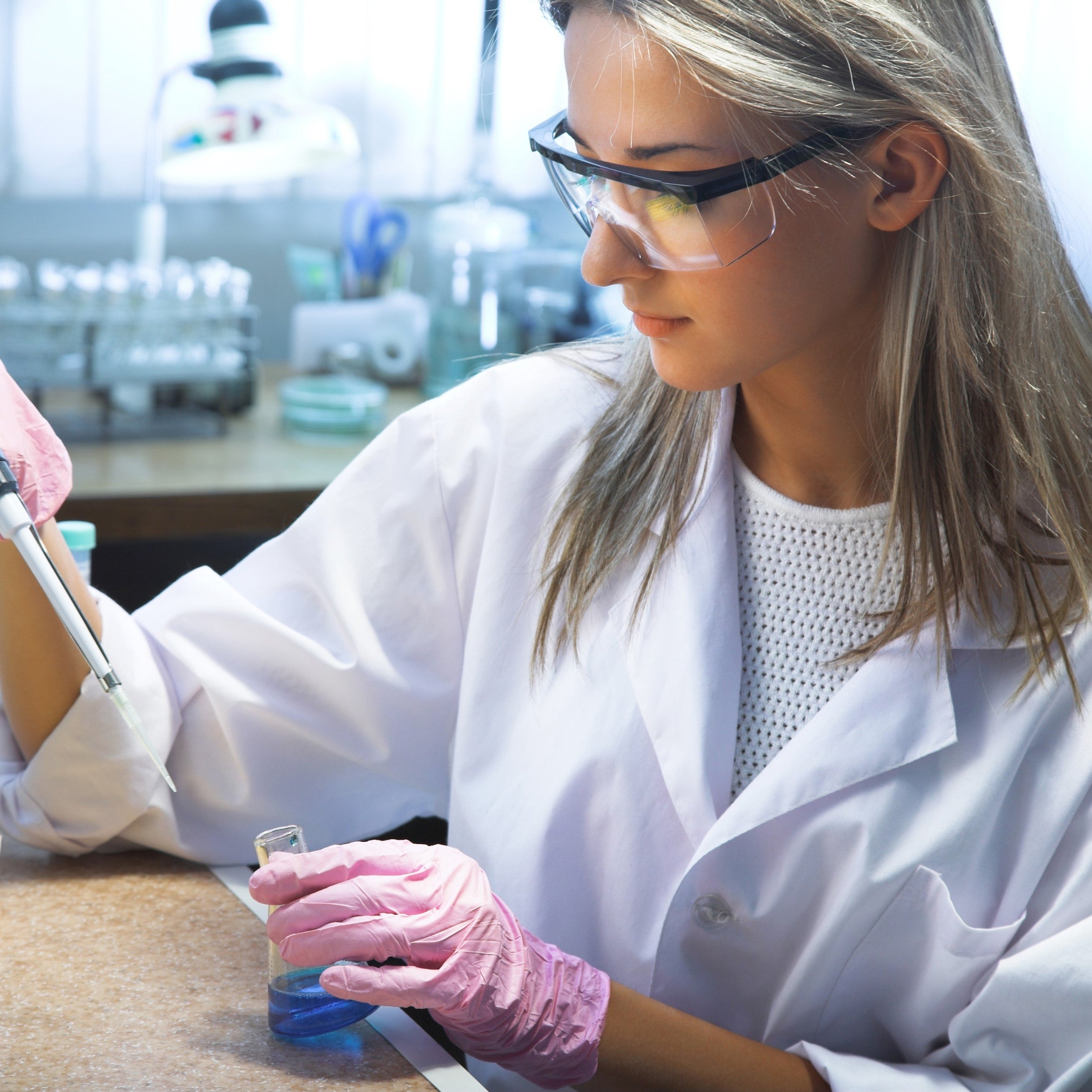 <Female lab analyst, wearing safety glasses and pink gloves, pipetting a liquid into a blue liquid in a small glass conical flask. Credit> AdobeStock_44142293