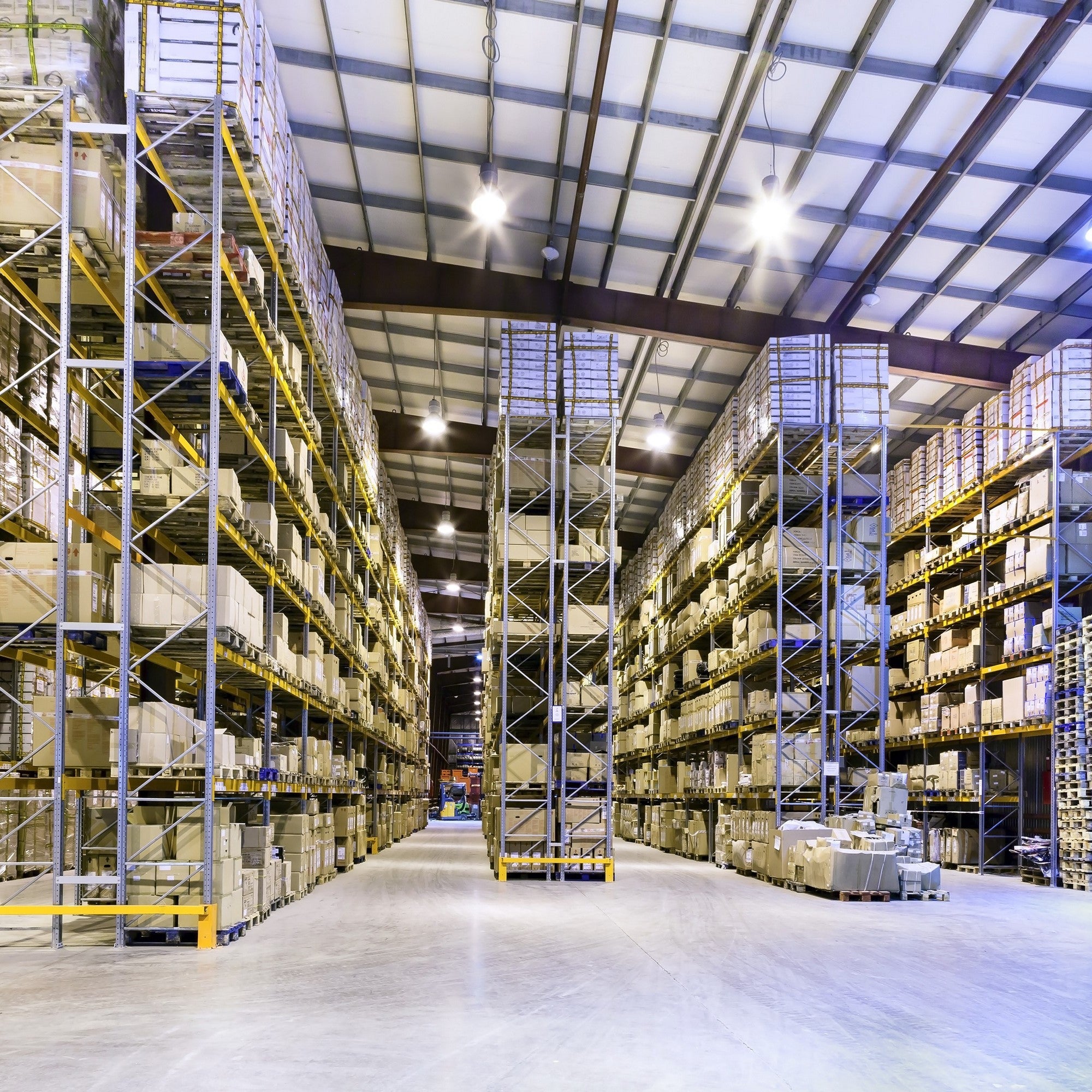 <Very clean and well organised warehouse with rows of racking holding pallets of boxes and grey floor under a sloping metal roof. Credit> AdobeStock_51465184