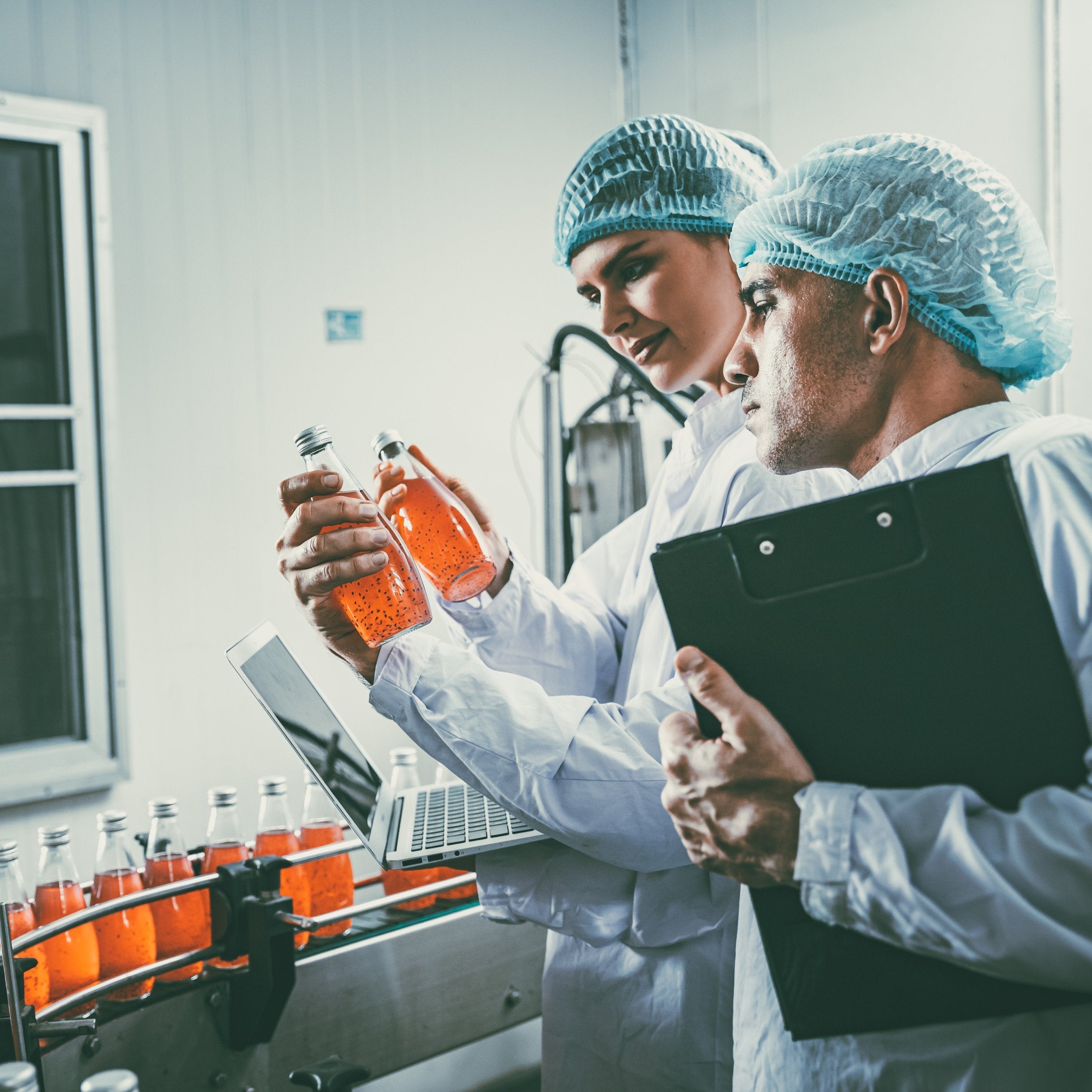 <Male and female quality officers checking bottles of orange liquid running down a conveyor belt on a production line. Credit> AdobeStock_552543492