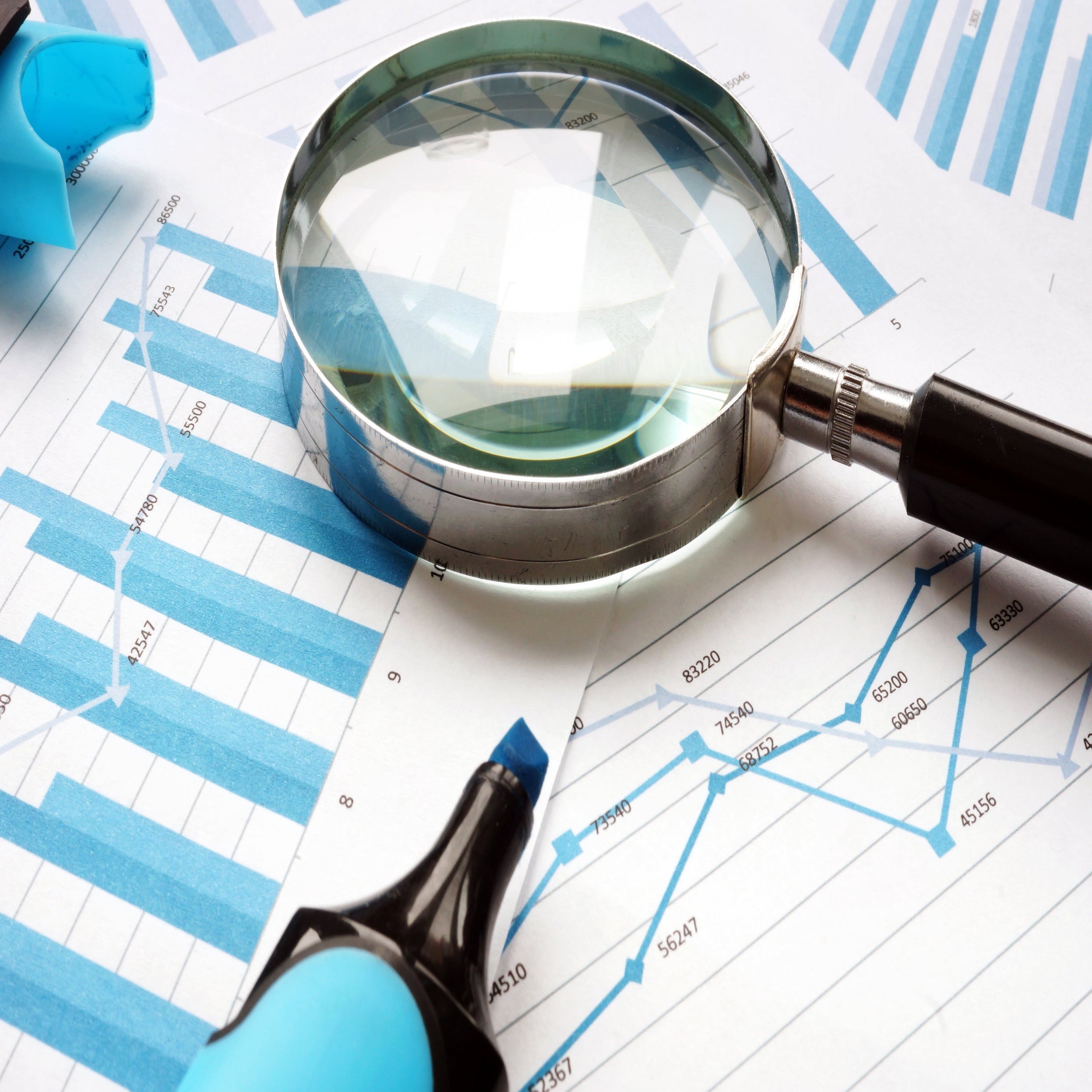 <Magnifying glass with black handle and chrome frame on top of blue graphs and bar charts of trends. Credit> 135091406 © Designer491 | Dreamstime.com