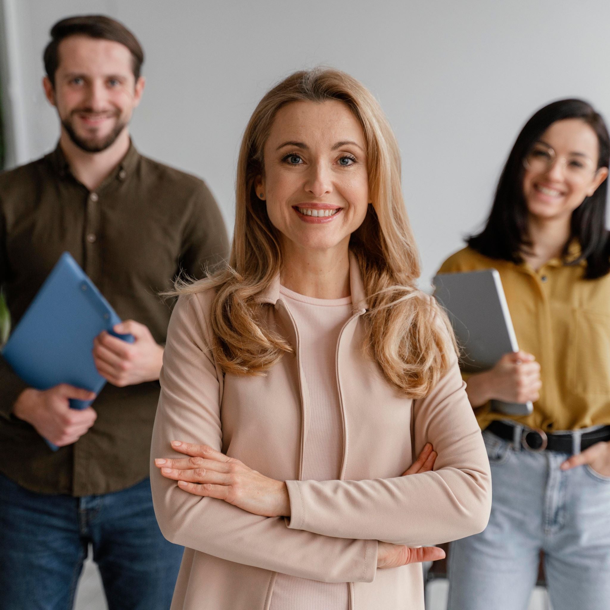 <Three smiling employees, a female at the front with her arms crossed, and a male and a female behind holding folders. Credit> Freepik