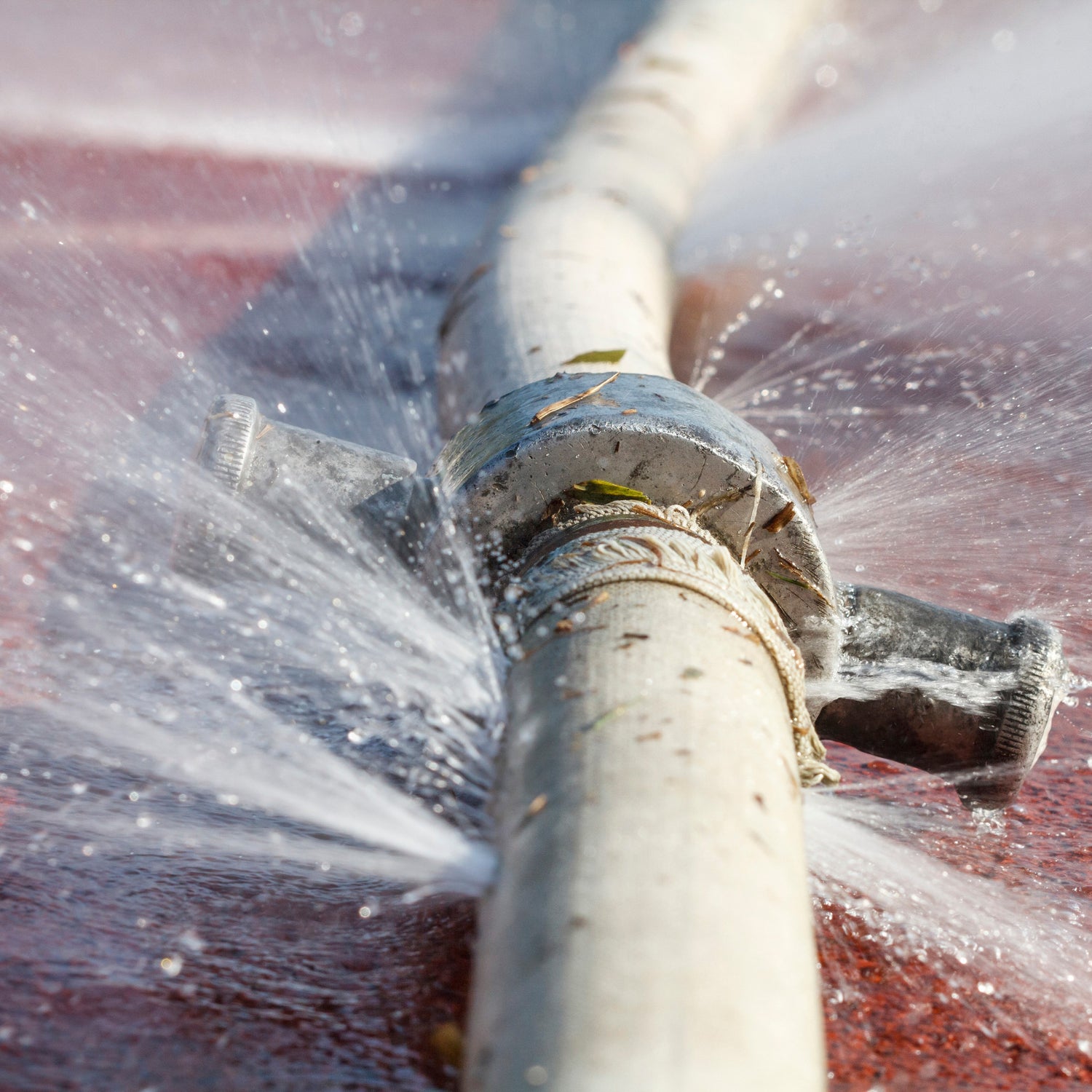 &lt;Burst, white, 10cm diameter water pipe or hose crossing a sunlit, red concrete surface  leaking water in several places under pressure. Credit&gt; AdobeStock_119492760