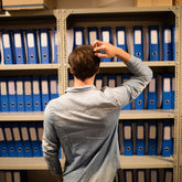 <Back of a male employee, wearing a denim coloured shirt, scratching his head trying to think in front of two tall shelf units full of blue A4 box folders. Credit> AdobeStock_146037976