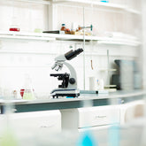 <White and black microscope on a white lab worktop with clean and modern white cupboards and drawers underneath and some samples in flasks on the shelf above. Credit> AdobeStock_158893280