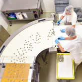 <Overhead view of two female production operators wearing white coats, hairnets and blue gloves, receiving dietary supplement lozenges from a wide, curved conveyor belt out of a cooling tunnel and placing them into metal trays for packing. Credit> AdobeStock_160608809