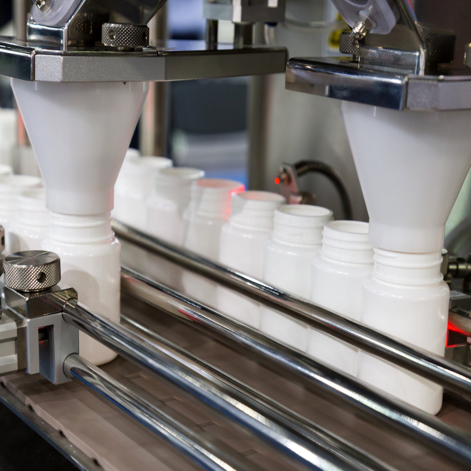 &lt;Two rows of white bottles passing down a conveyor belt, held in place with two chrome circular guardrails on each side, being filled with dietary supplement capsules from a white funnel connected to a filling machine. Credit&gt; AdobeStock_169195287