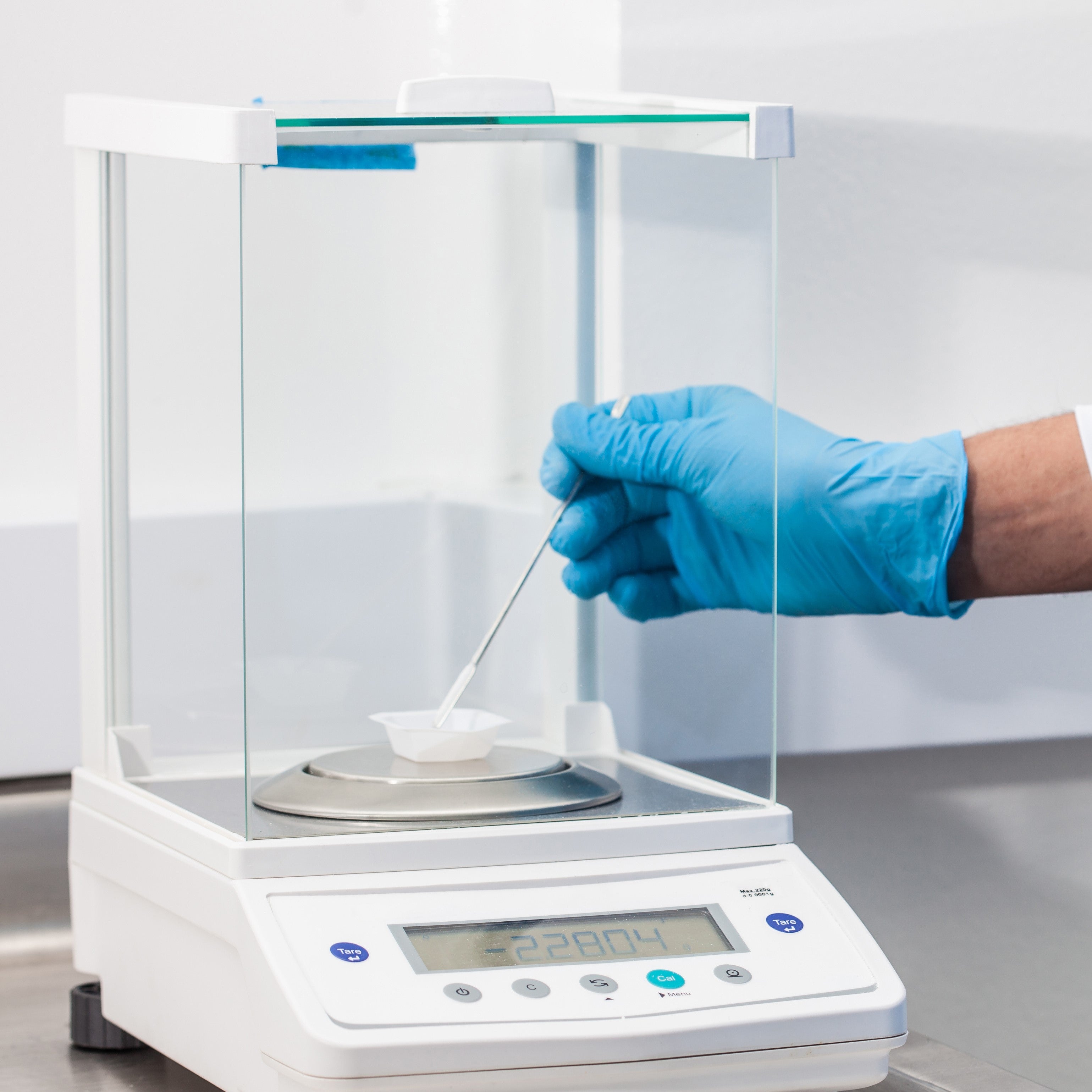 &lt;Male hand with blue glove adding powder with a spatula to a weigh boat on a white, digital analytical balance with clear glass screens. Credit&gt; AdobeStock_178852086