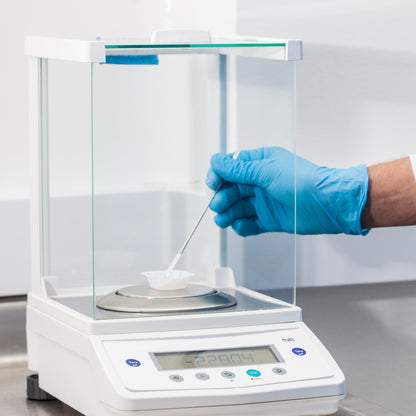 &lt;Male hand wearing a blue glove holding a spatula and using it to add more powder to a square plastic weigh boat on an analytical balance. Credit&gt; AdobeStock_178852086