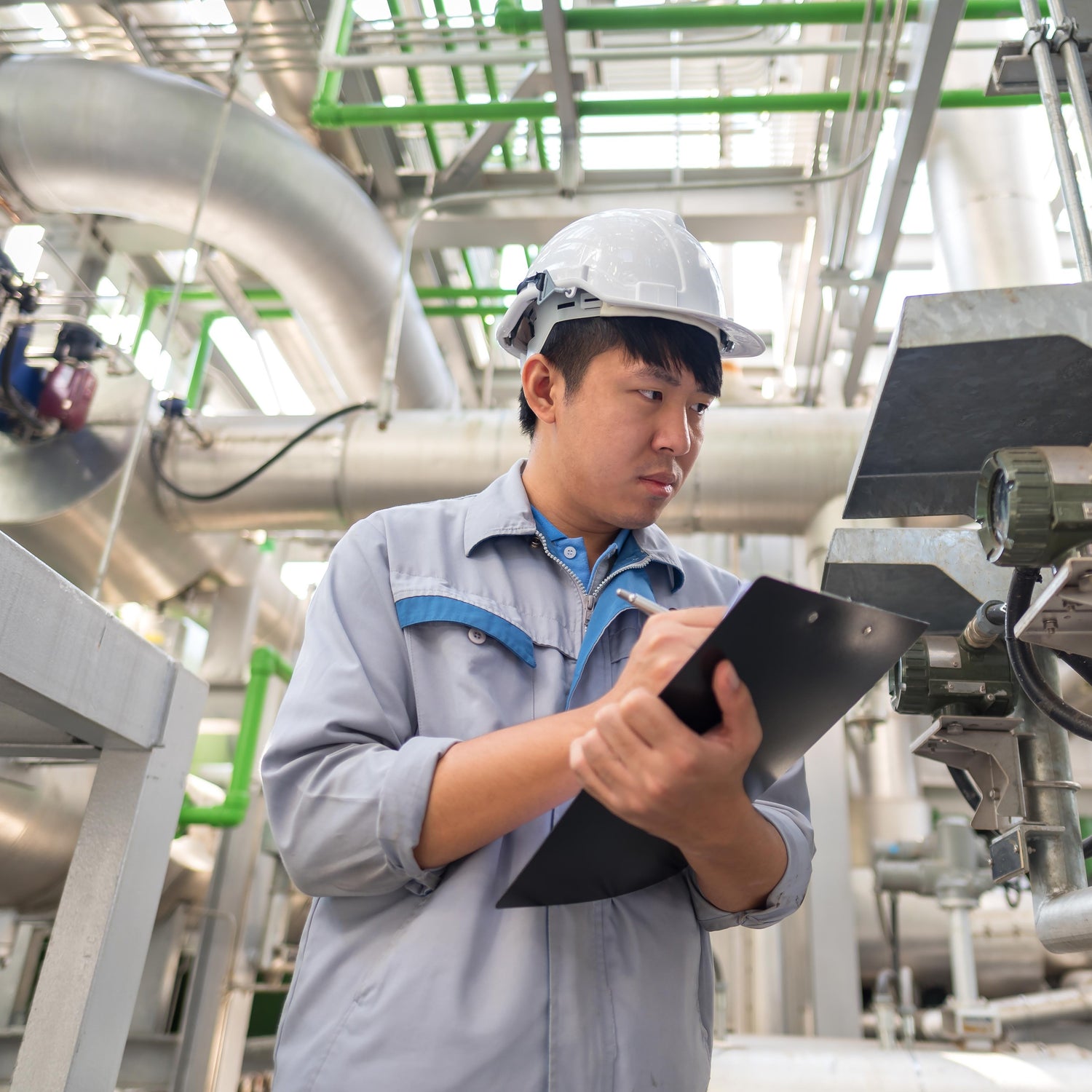 &lt;Male engineer, wearing grey overalls and white hard hat, stood in the production area surrounded by stainless steel pipework taking readings from a meter on a clipboard. Credit&gt; AdobeStock_196127416