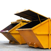 <Two large yellow skip bins with open lids sitting on a concrete yard next to a smooth white wall of a building. Credit> AdobeStock_203159028