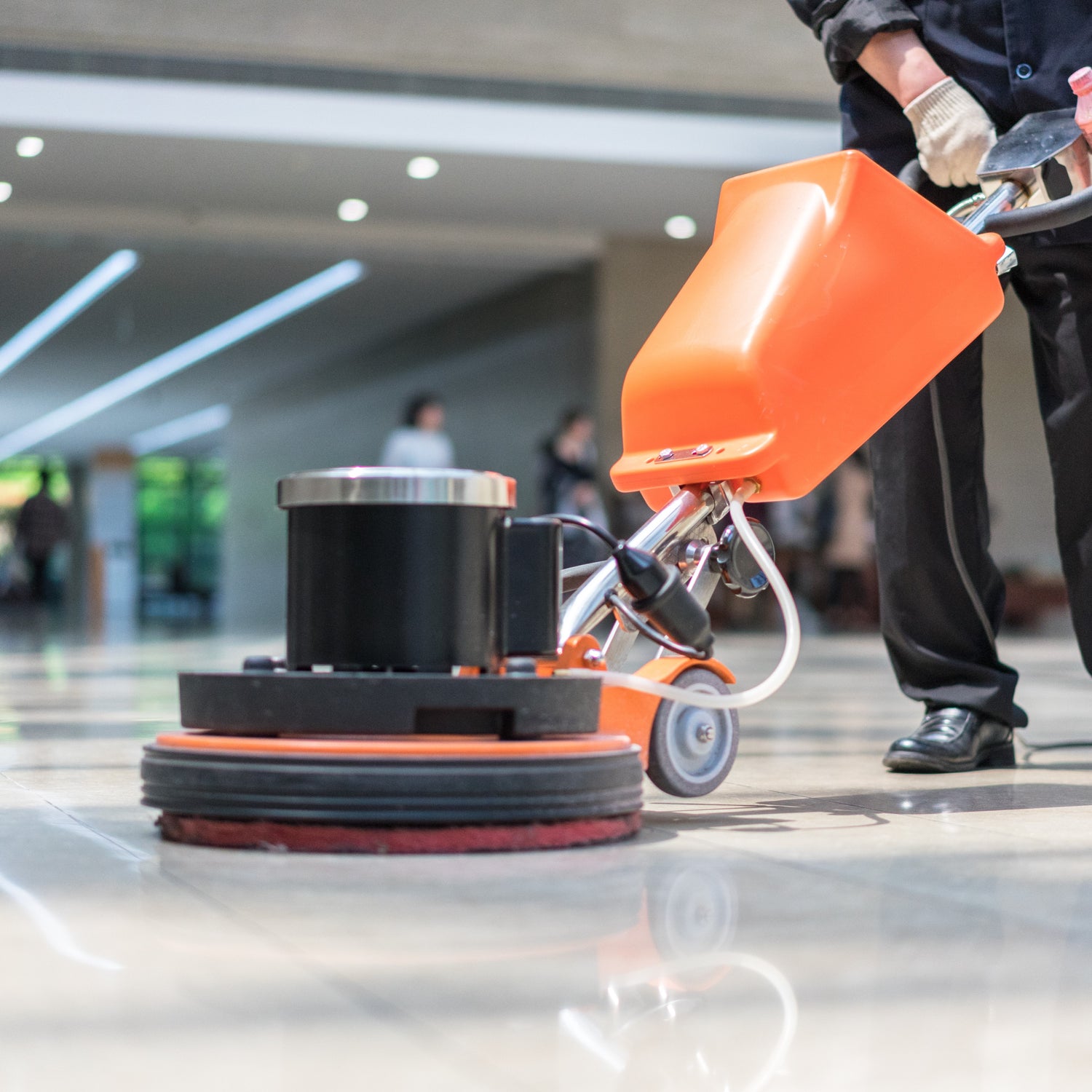 &lt;Legs of a man dressed in black uniform using a floor polisher on the cream marble floor of a floodlit office reception with employees blurred in the background entering the building. Credit&gt; AdobeStock_205886837