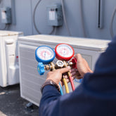 <Arm of a Maintenance Engineer wearing blue overalls checking a large white air conditioning unit in the outside yard with some pressure gauges. Credit> AdobeStock_210408504