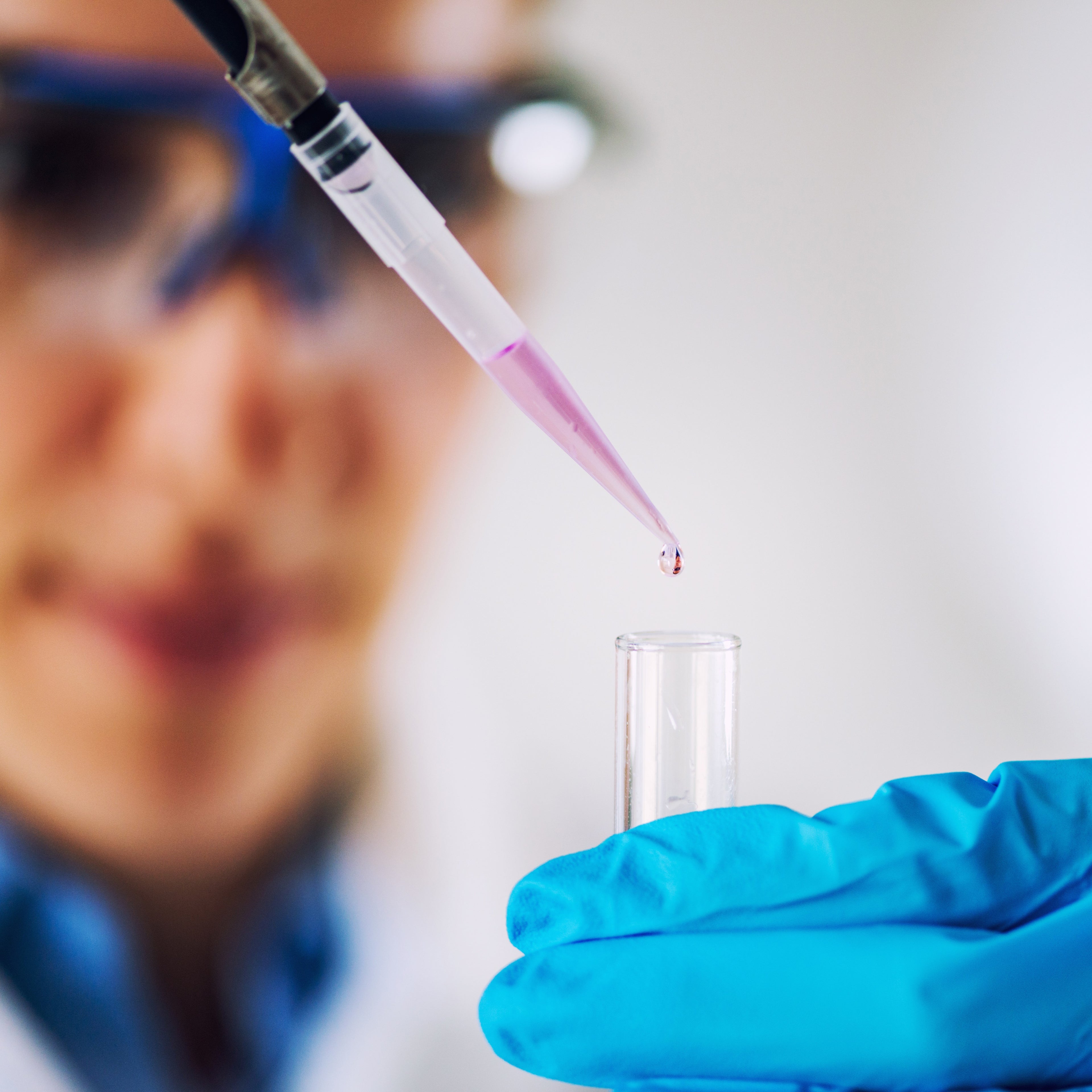 &lt;Blurred laboratory analyst wearing safety specs in background with close up of pipetting a clear, purple liquid into a test tube. Credit&gt; AdobeStock_212916043