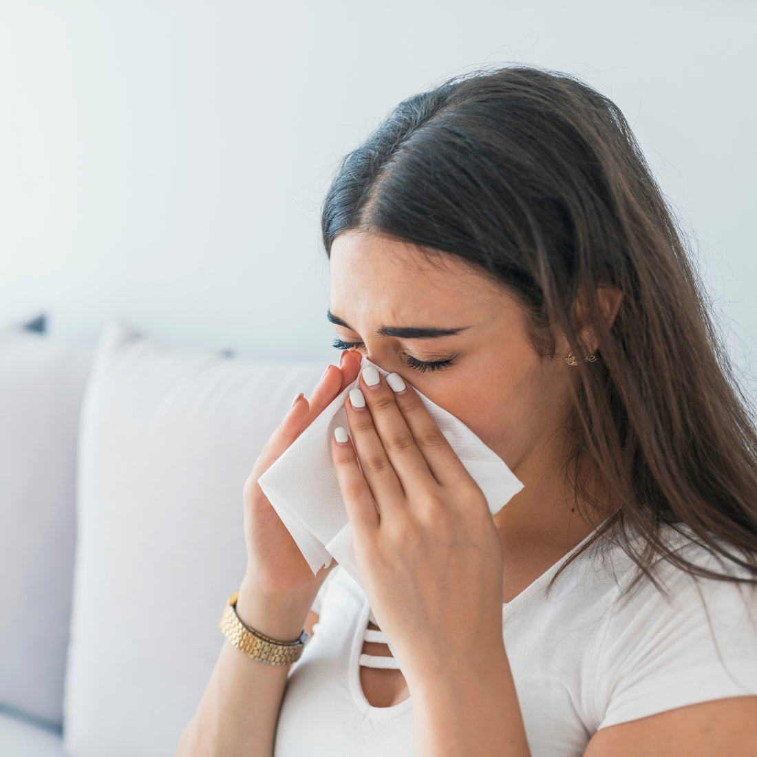 &lt;Brunette female employee, wearing a white t-shirt and gold watch, off work sick sitting on a white sofa at home blowing her nose into a tissue with cold and flu symptoms. Credit&gt; AdobeStock_219422085