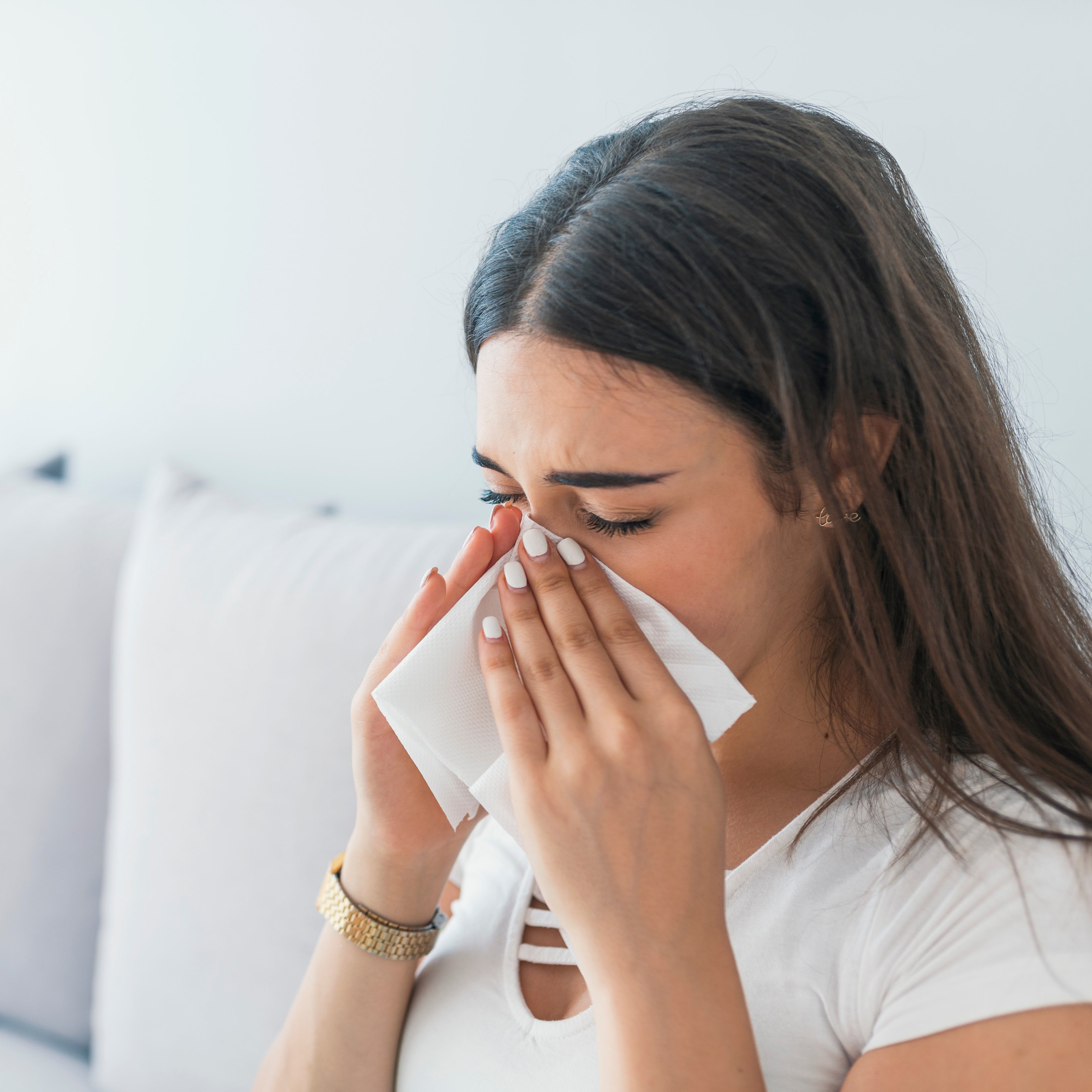 &lt;Brunette female employee, wearing a white t-shirt and gold watch, off work sick sitting on a white sofa at home blowing her nose into a tissue with cold and flu symptoms. Credit&gt; AdobeStock_219422085