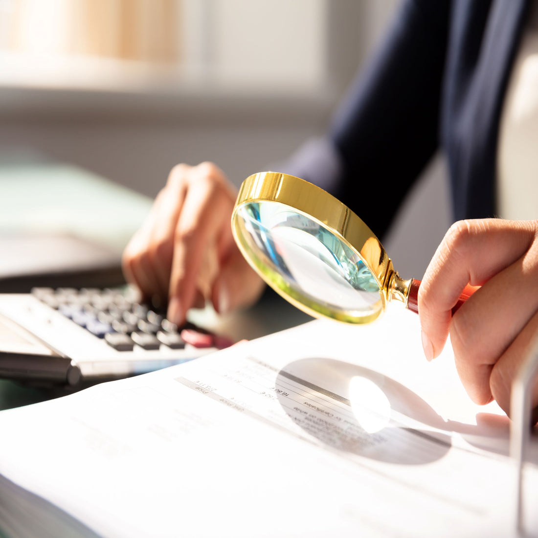 &lt;Arms of a woman holding a bright, gold magnifying glass in her left hand to review a document whilst typing on a laptop keyboard with her right hand. Credit&gt; AdobeStock_224910698