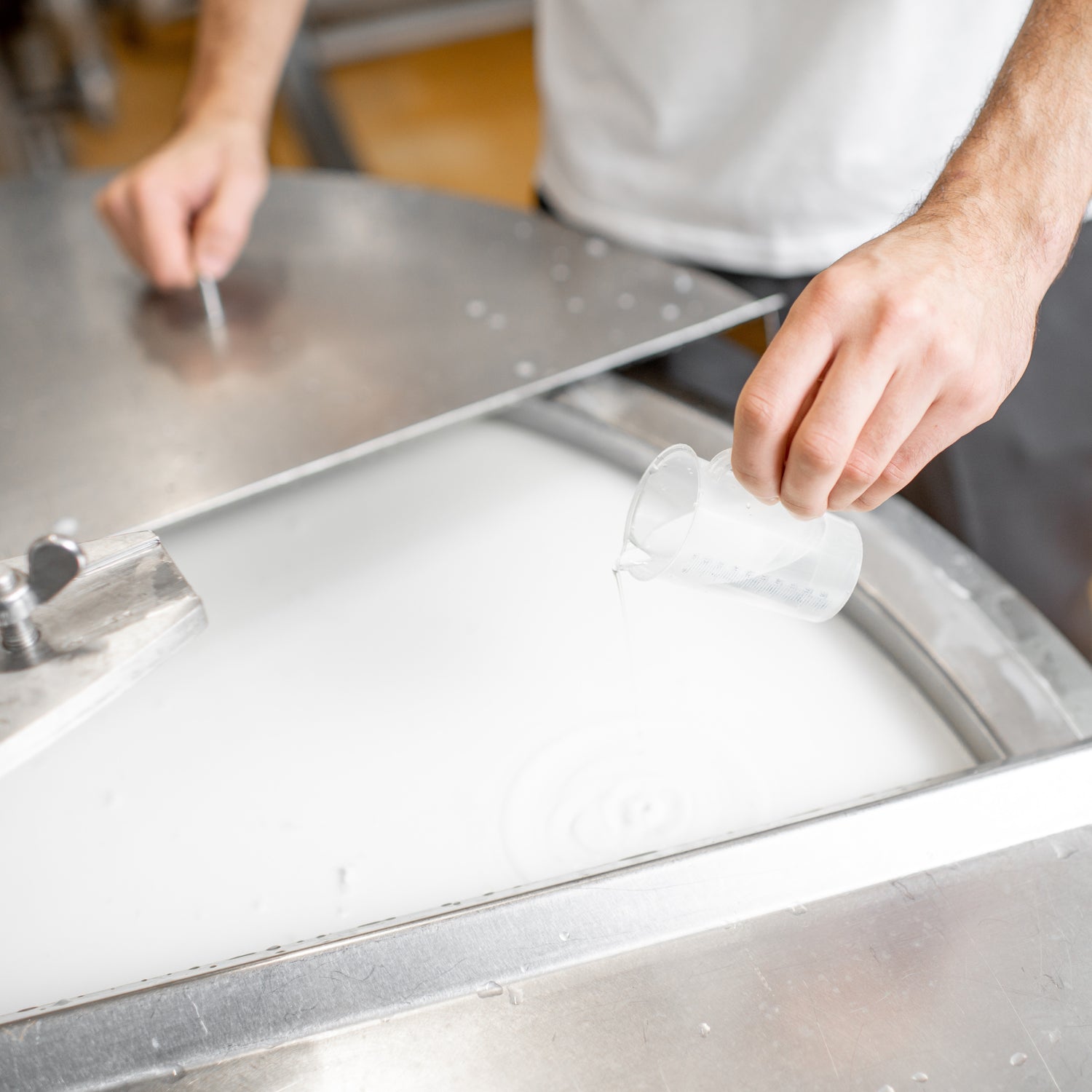 &lt;Body of manufacturing operator wearing white overall and grey trousers, pouring a clear liquid from a small plastic jug into a batch of cream in a mixing vessel. Credit&gt; AdobeStock_235550619