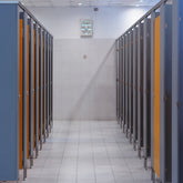 <Employee series of blue grey toilet cubicles with wooden doors in a rest area with grey walls, white tiled floors and white ceiling grid with wall ventilation fan. Credit> AdobeStock_239768281