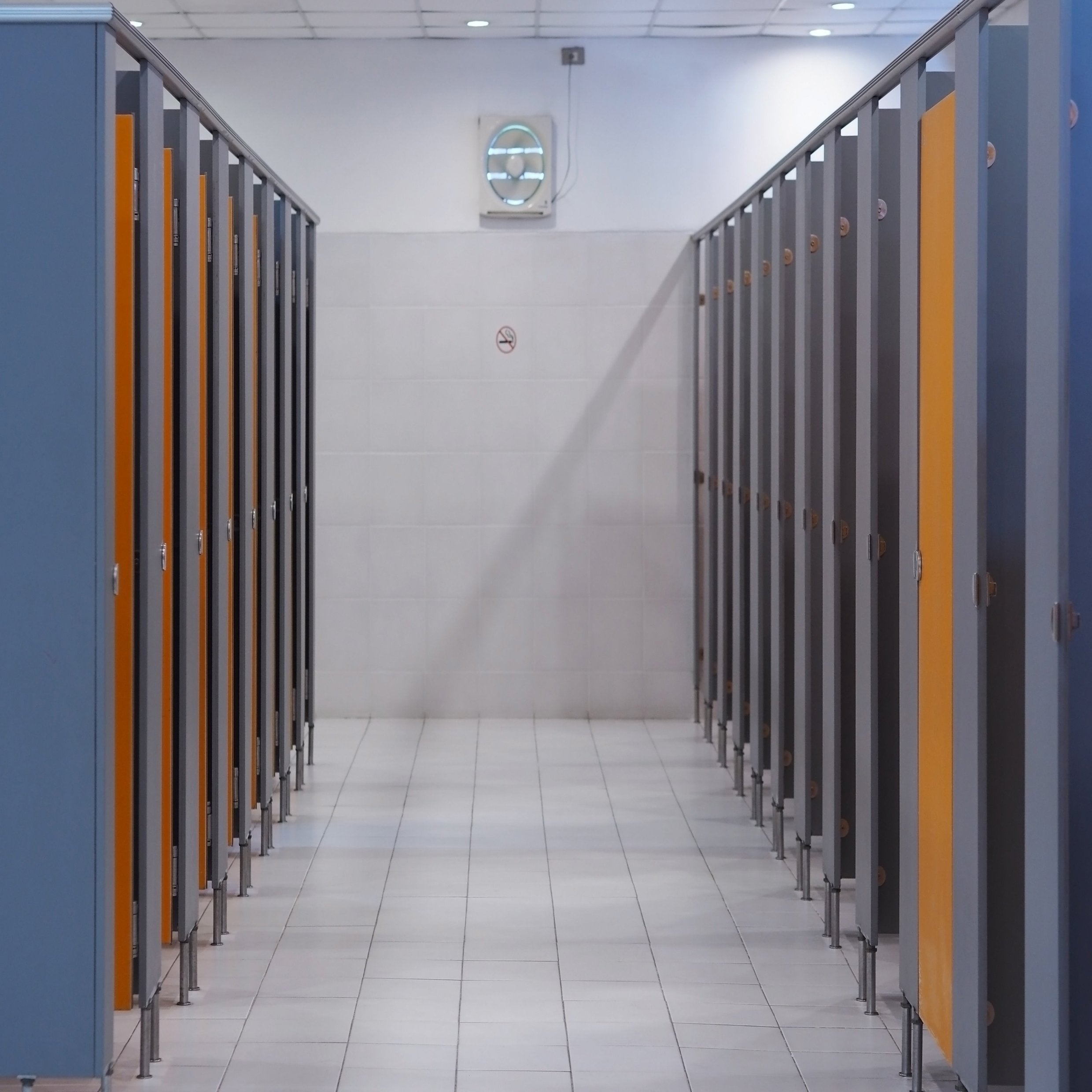 &lt;Employee series of blue grey toilet cubicles with wooden doors in a rest area with grey walls, white tiled floors and white ceiling grid with wall ventilation fan. Credit&gt; AdobeStock_239768281