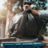 <Arm of a man wearing light blue overalls putting a black bin bag of food waste into a turquoise wheelie bin in front of a concrete wall with a tree in the blurred background. Credit> AdobeStock_240623500