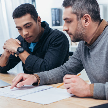 &lt;Two male supervisors, wearing navy blue and grey jumpers,  sat at a wooden office desk reviewing production paperwork, one of them pointing at something in the documents. Credit&gt; AdobeStock_241546209