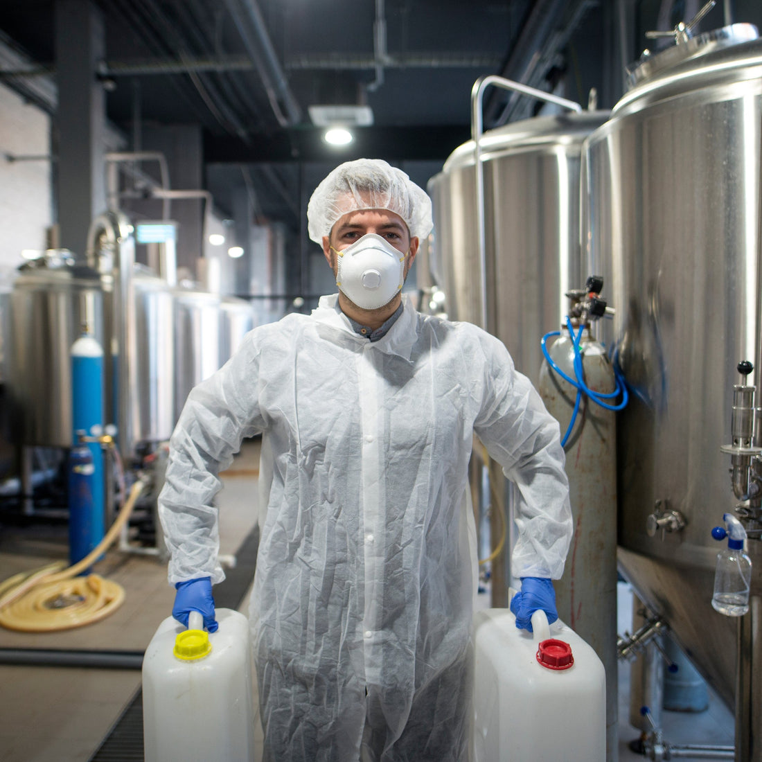 &lt;Male production operator wearing white overcoat, hairnet, face mask and blue gloves, in a manufacturing unit with several mixing tanks, carrying two jerry cans with cleaning liquids with yellow and red caps. Credit&gt; AdobeStock_244374945