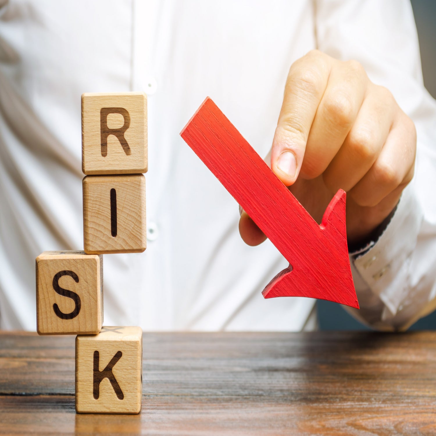 &lt;Body and arm of a man, wearing a white shirt, sat at a wooden table on which four wooden blocks are stacked to spell Risk where the S is almost falling, and his finger is on a red arrow pointing downwards to minimize risk. Credit&gt; AdobeStock_270610352