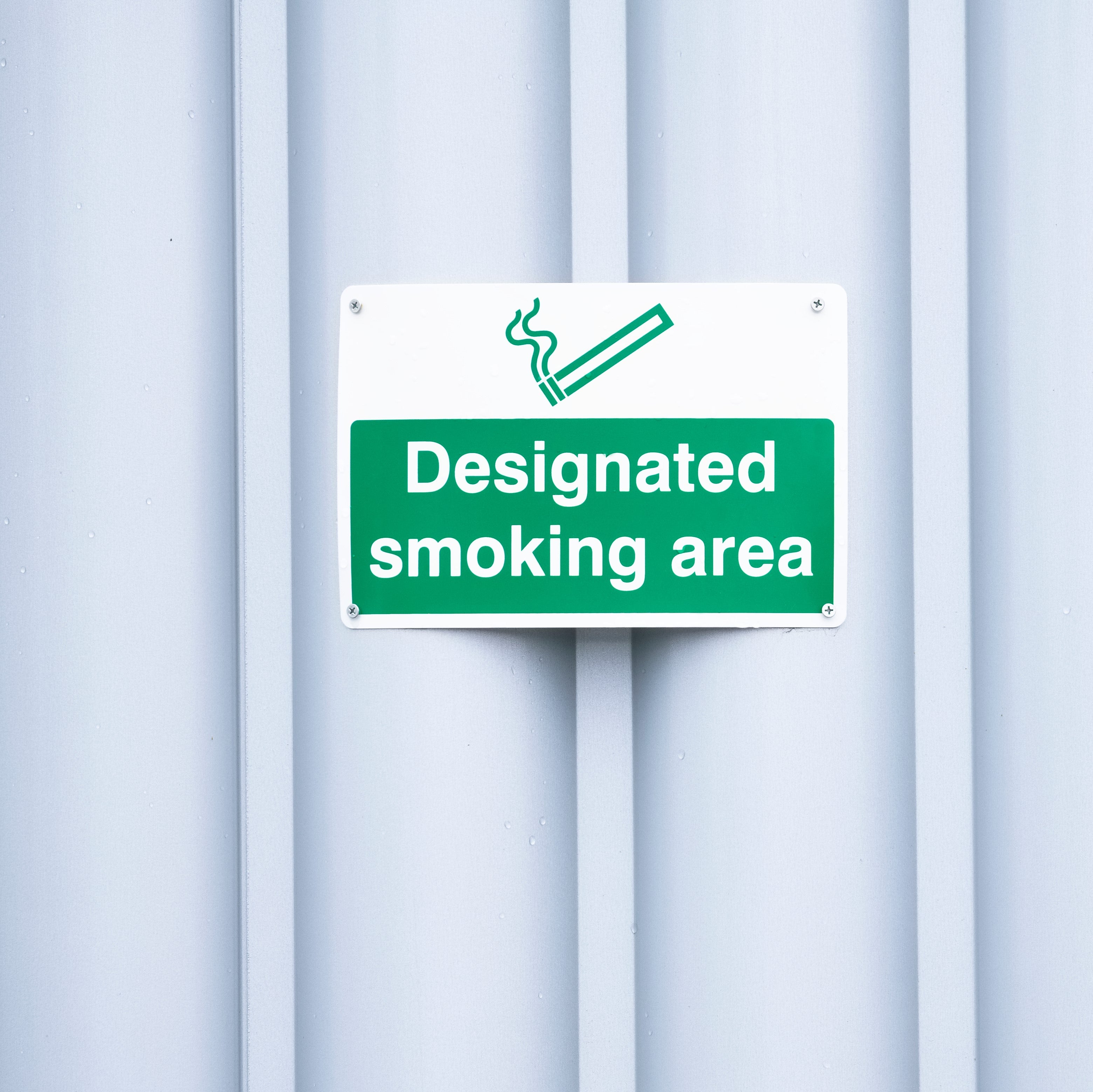 &lt;Green sign on a white corrugated metal building to direct personnel to a designated smoking area on site. Credit&gt; AdobeStock_272303046