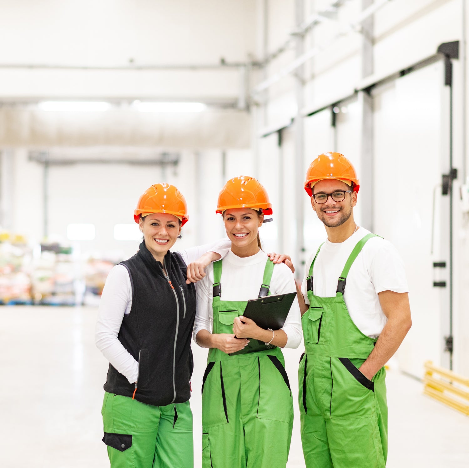 &lt;Three smiling production engineers, two female and one male, wearing green overalls and orange hard hats stood in a white clean production area. Credit&gt; AdobeStock_274216864