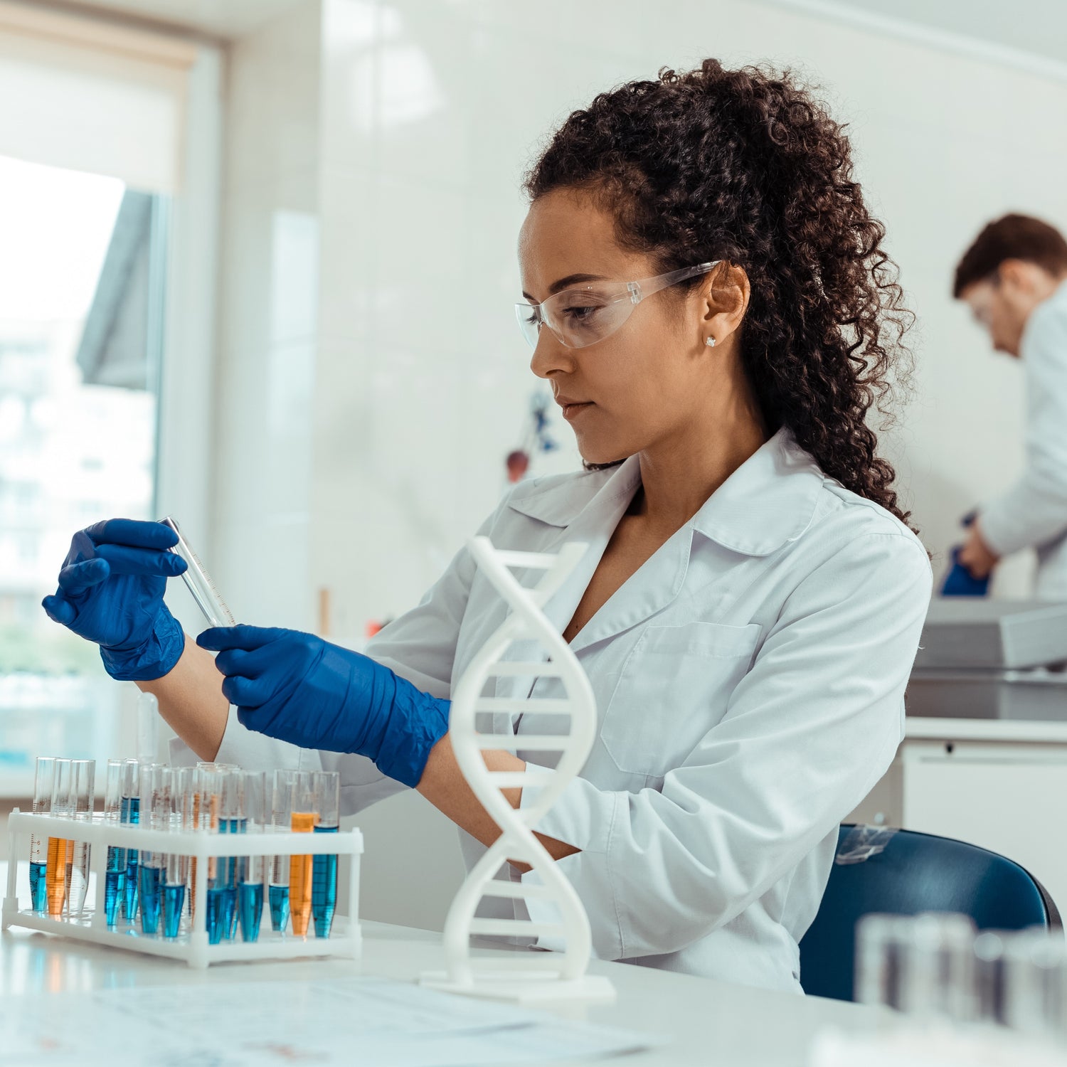 &lt;Female laboratory analyst weraing white coat and blue gloves, with male analyst in the blurred background, holding a glass test tube taken from a white rack  with 20 more with light blue or yellow liquid. Credit&gt; AdobeStock_275088574