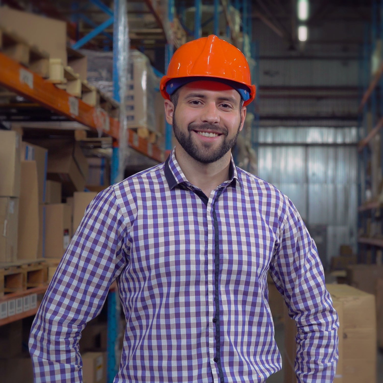 &lt;Smiling, male training supervisor in the warehouse stood in fron tof racking with boxes, wearing a blue checked shirt and orange hard hat. Credit&gt; AdobeStock_277436381