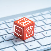 <Red cube with emergency e-mail, letter, and phone symbols on the three visible faces, sitting on a white laptop keyboard. Credit> AdobeStock_277649030