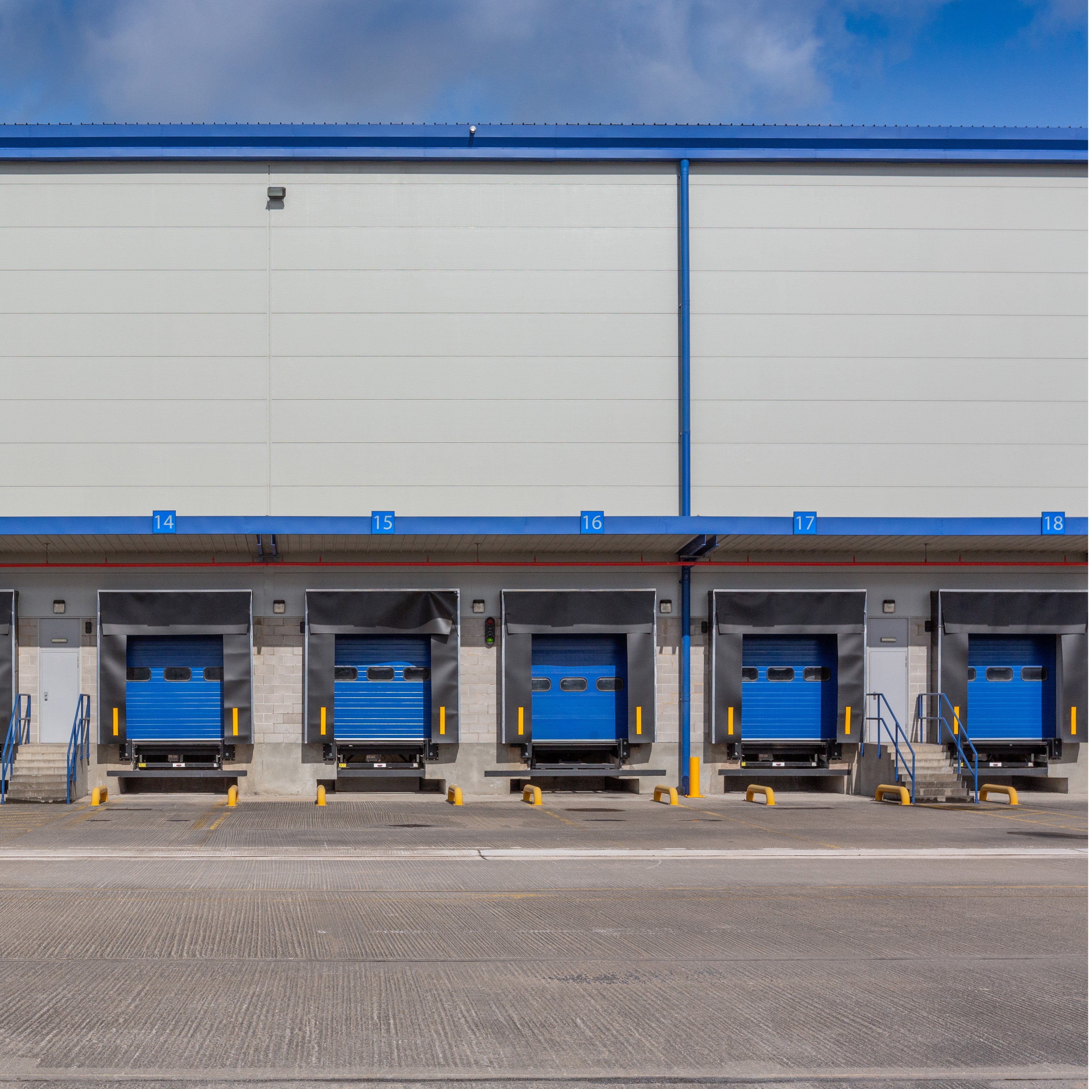 &lt;View from the outside concrete yard in sunshine of five blue roller shutter dock doors for loading and unloading lorries into a grey metal building numbered 14 to 18. Credit&gt; AdobeStock_292350584