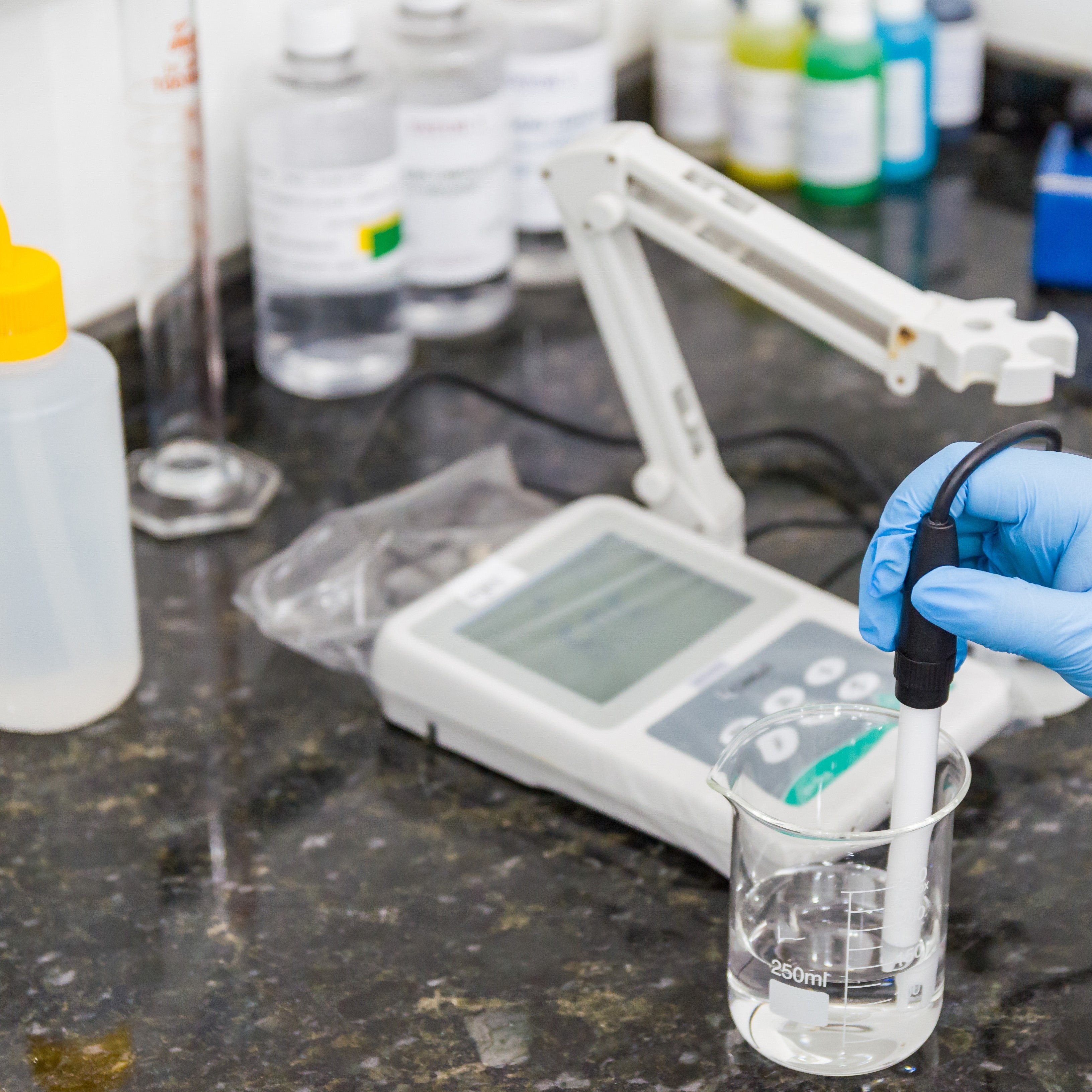 &lt;Hand in a blue glove holding the probe of a pH meter in a glass beaker of clear liquid, on a stone effect worktop, with bottles of buffer solution. Credit&gt; AdobeStock_296409151