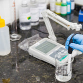 <Hand wearing a blue glove holding a pH probe in a glass beaker with clear liquid attached to a pH meter. Credit> AdobeStock_296409151