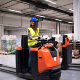 <Smiling, female warehouse operator with yellow fluorescent jacket and blue hard hat driving an orange electric forklift pallet truck in a warehouse near wooden pallets of shrink-wrapped boxes. Credit> AdobeStock_302493850