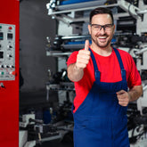 <Maintenance Engineer wearing red T-shirt, blue overalls and safety glasses giving a thumbs up sign that the laminate filling machine behind him and the red control panel next to him is working. Credit> AdobeStock_308218710