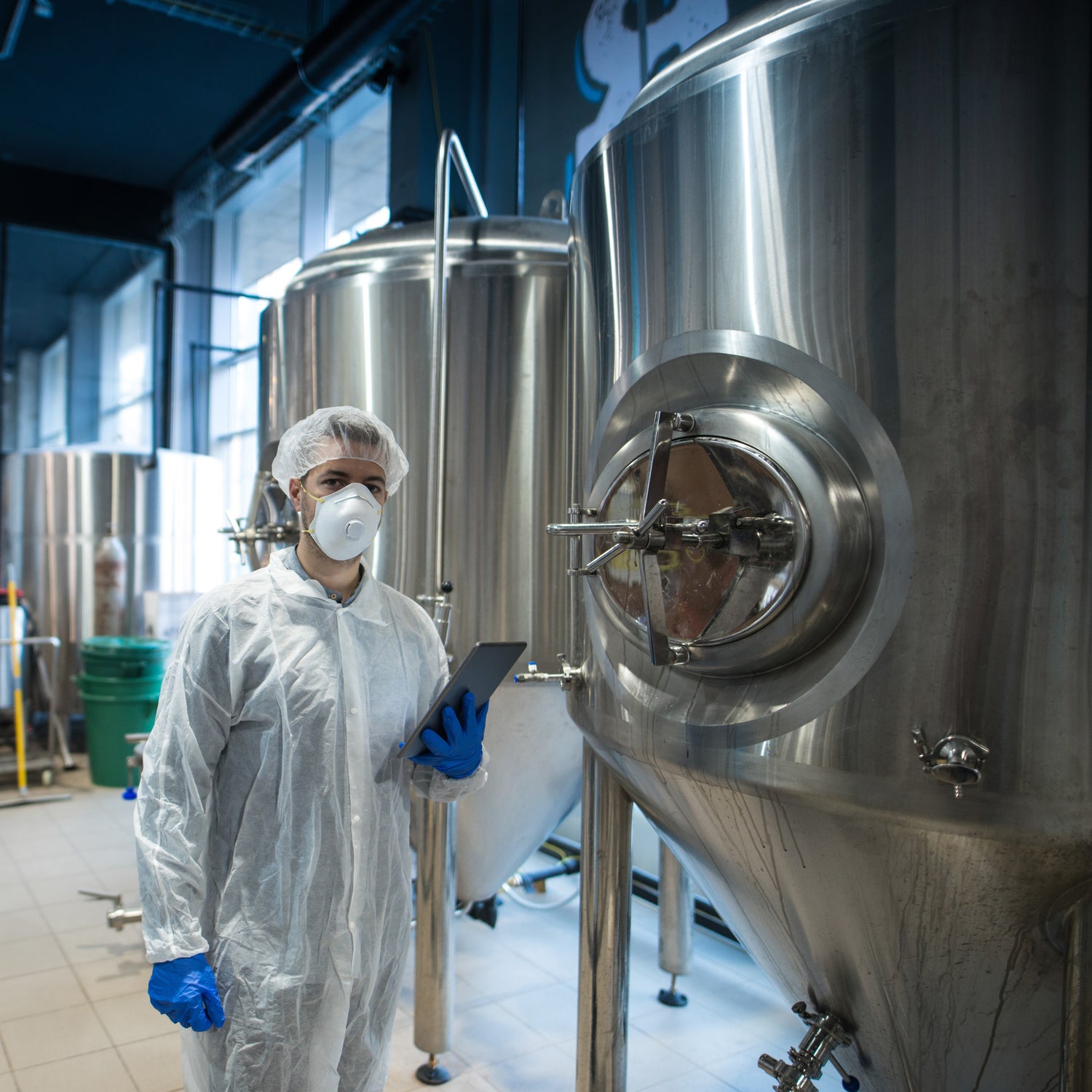 &lt;Male operator in white overalls, blue gloves and dust mask walking past a row of three stainless steel mixing tanks holding a tablet. Credit&gt; AdobeStock_314233485