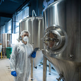 <Male operator in white overalls, blue gloves and dust mask walking past a row of three stainless steel mixing tanks holding a tablet. Credit> AdobeStock_314233485