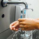 <Employee washing hands from an automated hands free chrome spout in a grey plastic trough next to a white liquid soap dispenser. Credit> AdobeStock_315036523