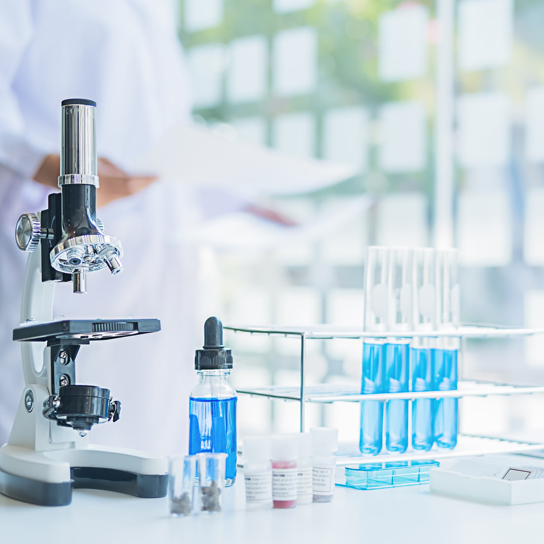 &lt;White, black, and chrome microscope on a  white lab surface next to a rack with 4 test tubes of blue liquid and a lab analyst in white labcoat in a blurred background with  windows and green outside area. Credit&gt; AdobeStock_336473215