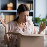 <Woman customer in cream top, sitting on a white sofa in the living room, open arms and mouth  in horror as she opens her box of dietary supplements that she ordered. Credit> AdobeStock_345250156