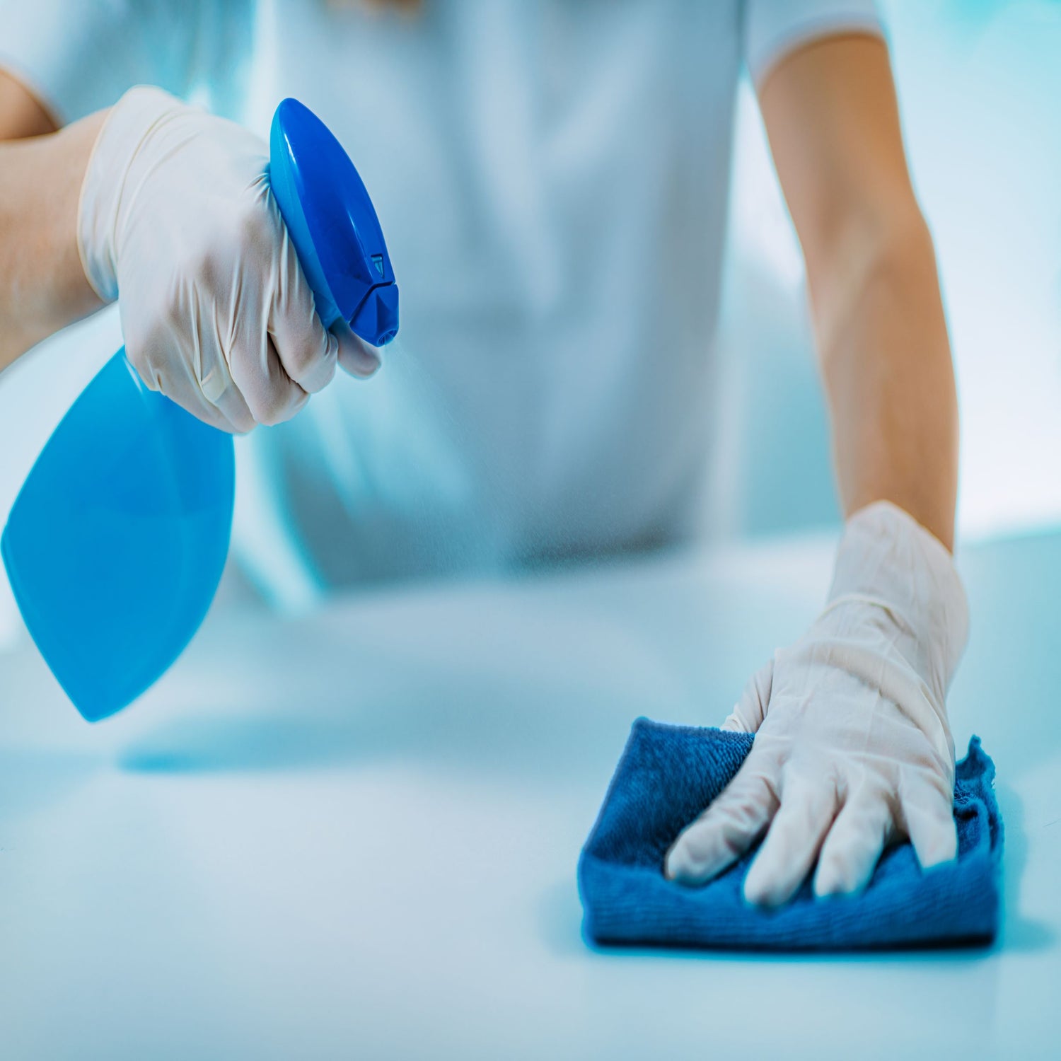 &lt;Arms and body of a woman wearing light blue overall and white gloves cleaning a white contact surface with a clean blue cloth and an approved  cleaning spray from a blue spray gun. Credit&gt; AdobeStock_346099456