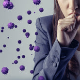 <Part of a face of a woman with long brown hair, wearing a grey blazer and white shirt, sneezing into her hand and releasing many purple, spherical, spiky virus particles into the air. Credit> AdobeStock_351167737