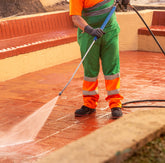 <Legs and body of a man wearing fluorescent green and orange workwear and safety boots pressure washing an employee outside tiled break area next to a tree and garden. Credit> AdobeStock_359884284