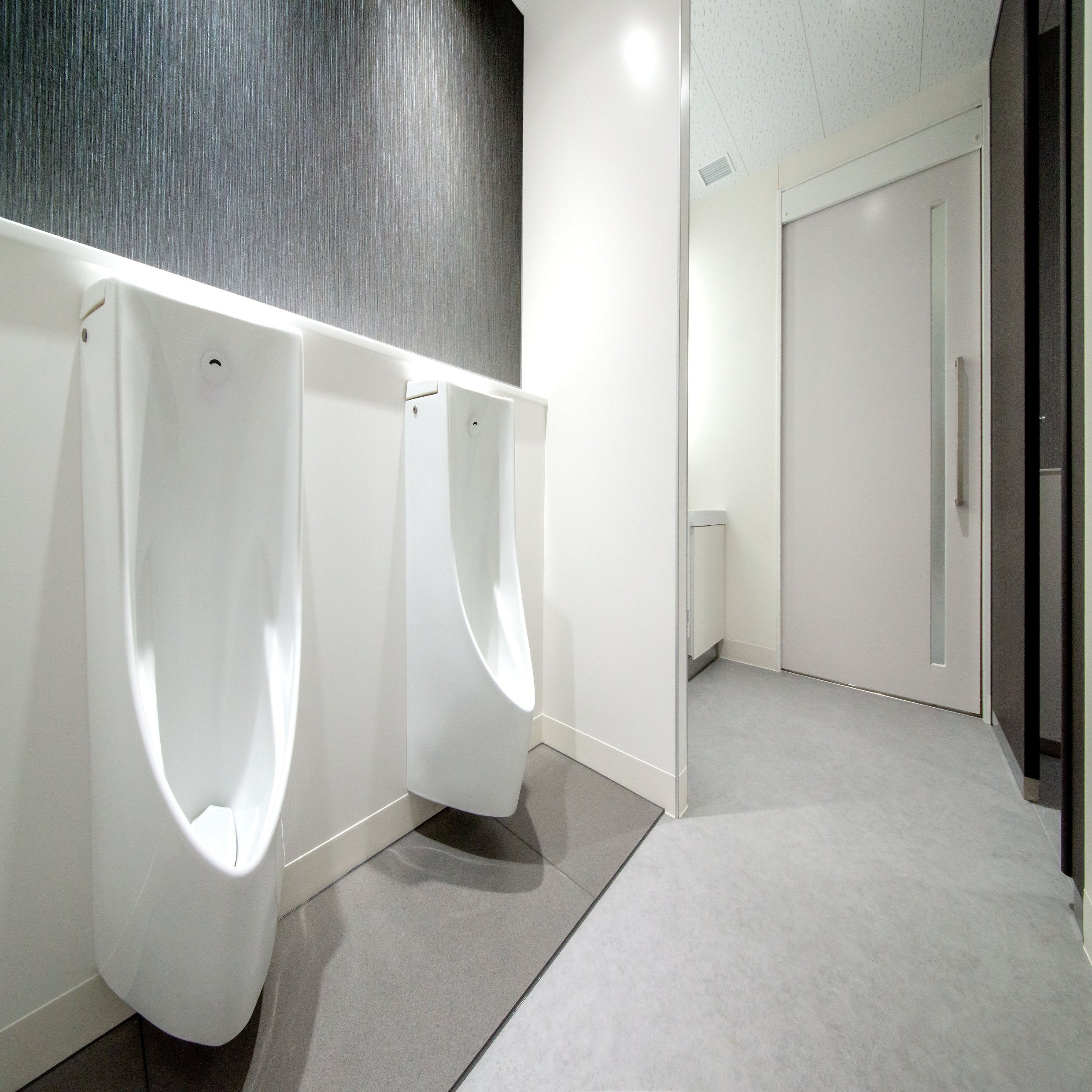 &lt;Two white urinals in an employee rest area with dark grey stone effect wall, grey vinyl floor and white walls and doors. Credit&gt; AdobeStock_364201756