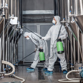 <Two male operators in full white overalls, face masks and blue overshoes, spraying cleaning fluid from green portable tanks on a series of stainless steel mixing tanks. Credit> AdobeStock_376451842