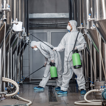 &lt;Two male operators in full white overalls, face masks and blue overshoes, spraying cleaning fluid from green portable tanks on a series of stainless steel mixing tanks. Credit&gt; AdobeStock_376451842